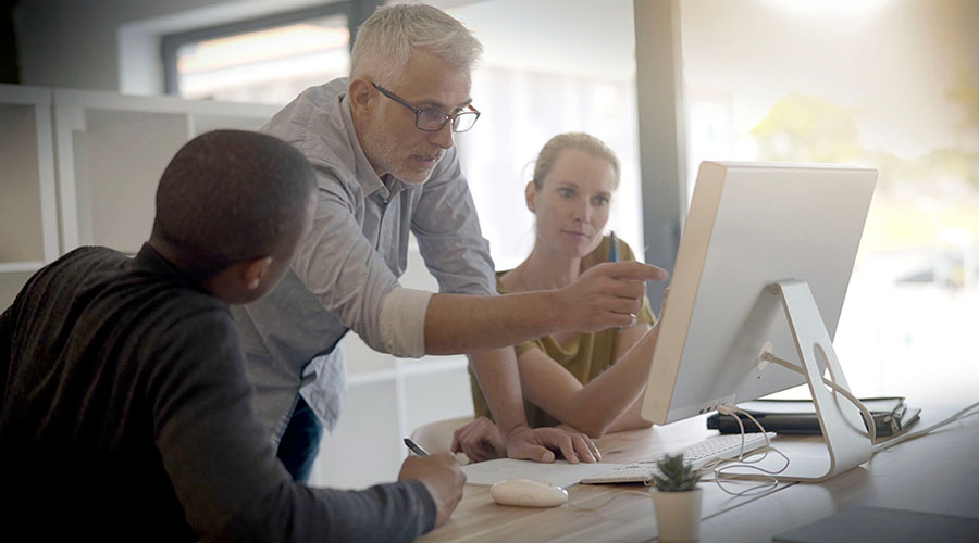 A man in a dress shirt pointing to a computer monitor while a man in a black long-sleeved shirt and a woman in a brown shirt are looking at the monitor.