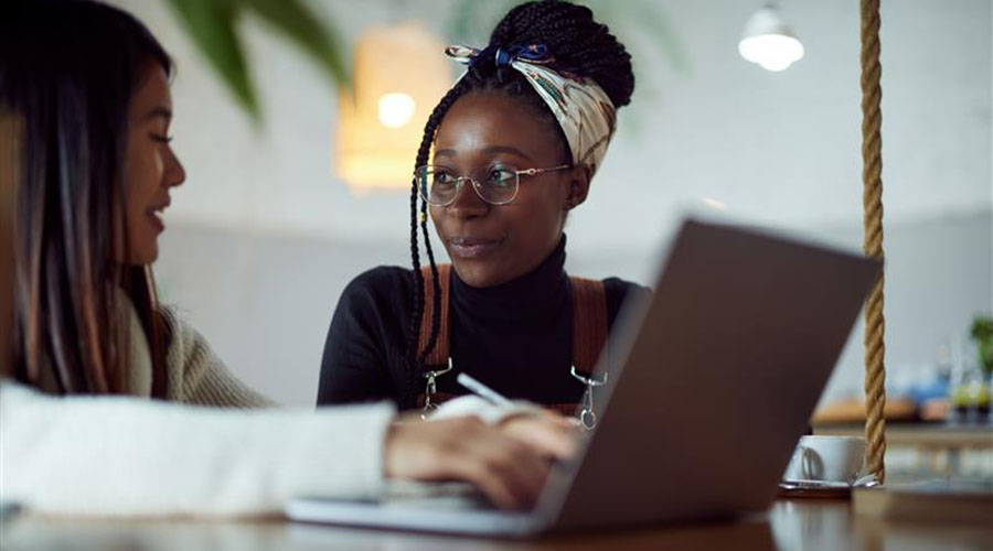 Woman in a black shirt sitting and looking at a girl in a white shirt, who is using a laptop.