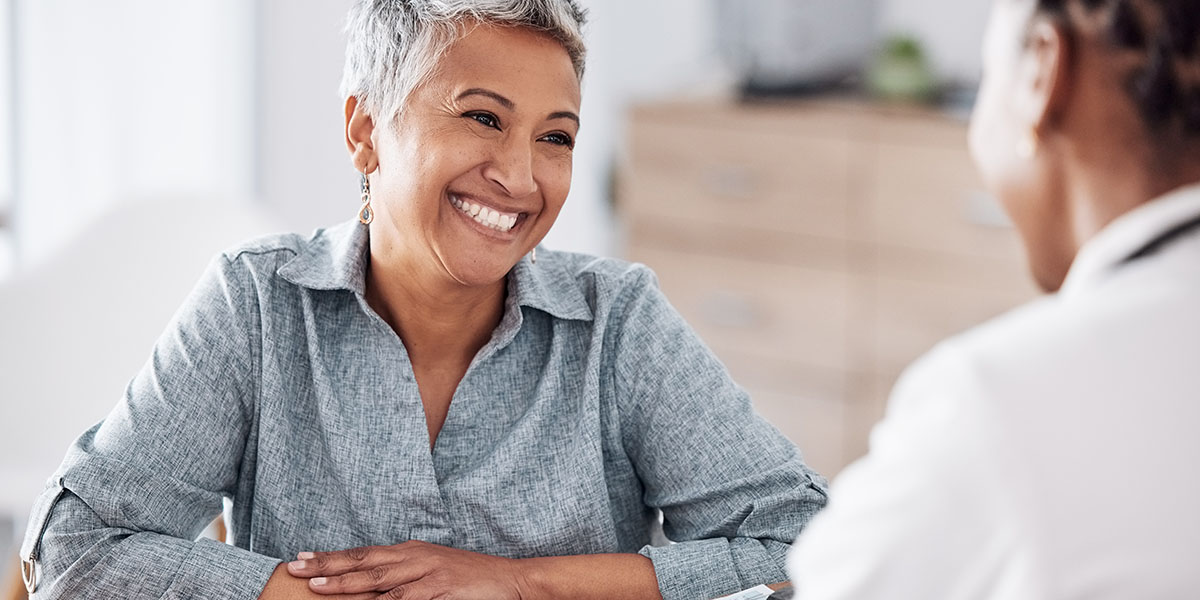 A woman with short gray hair is sitting at a desk, smiling at a healthcare professional in an office.