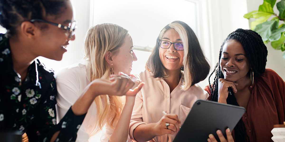 A diverse group of four women is gathered around a table, smiling and engaged in a discussion while one woman holds a tablet. 