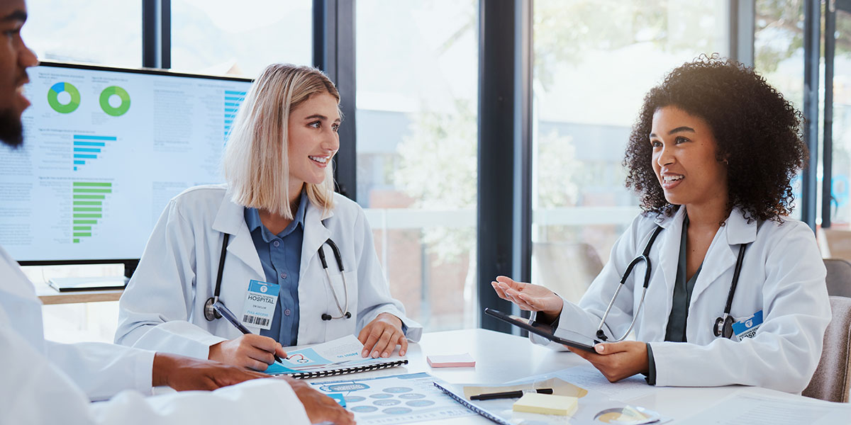 A group of healthcare professionals in lab coats are having a discussion around a table filled with documents and charts. 